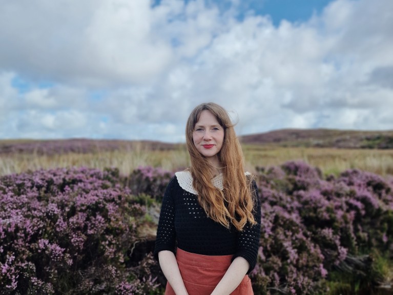 A blonde woman stands in front of a flowering hedge, the scenic Irish landscape lies beyond.