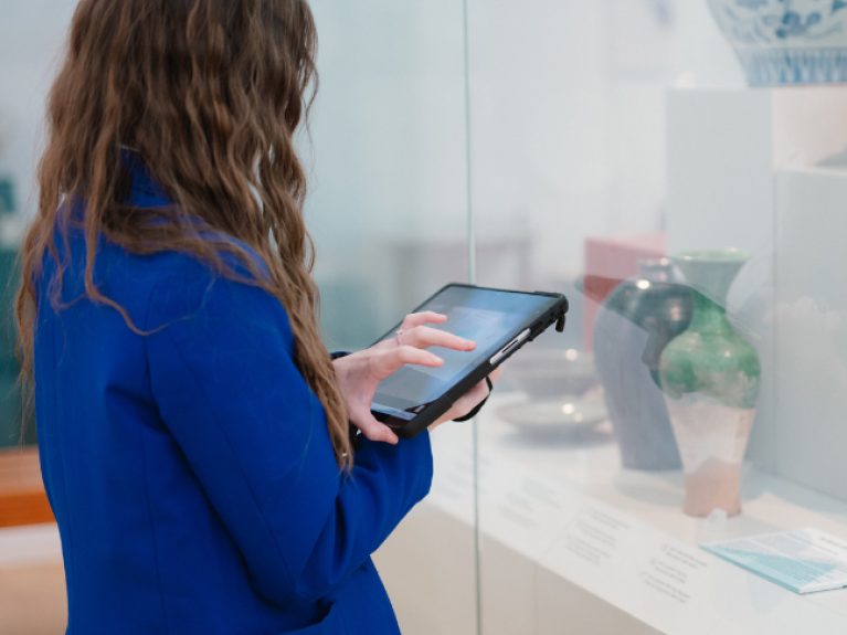 A school pupil wearing a blue blazer, holding an iPad, looking into a glass cabinet of ceramics in Ulster Museum.