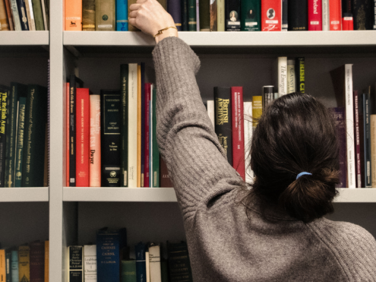 Woman with beige jumper and brown hair, tied up, reaching for a book from a book case