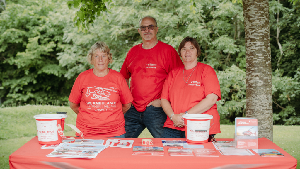 Table with a red table cloth and Air Ambulance NI branded collection buckets (to collect money), with three people standing behind the table dressed in red t-shirts for the charity.