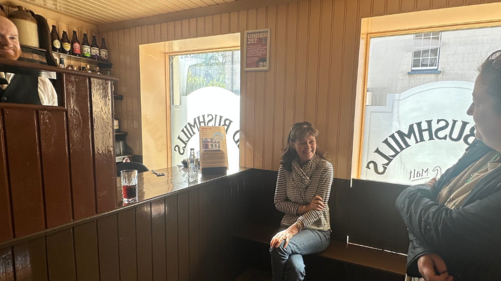 A woman sits on the bench of a pub. She is smiling at a second woman who is half in frame.