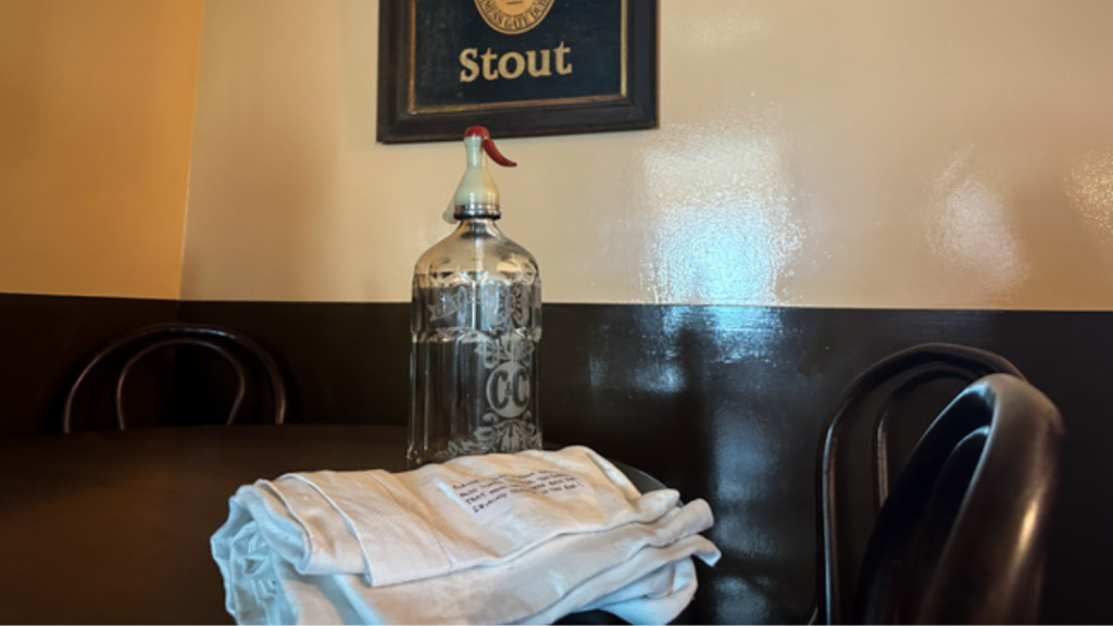 On a pub table, an old seltzer bottle and a pile of folded towels. 
