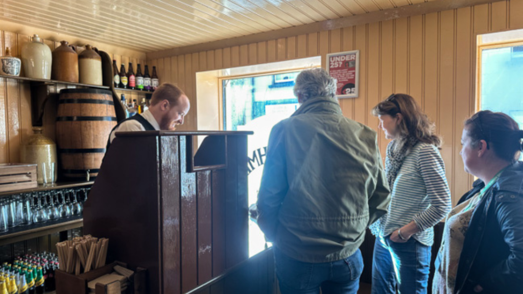 A group of people stand in an old fashioned pub. They have their backs to the camera, speaking to the barman.
