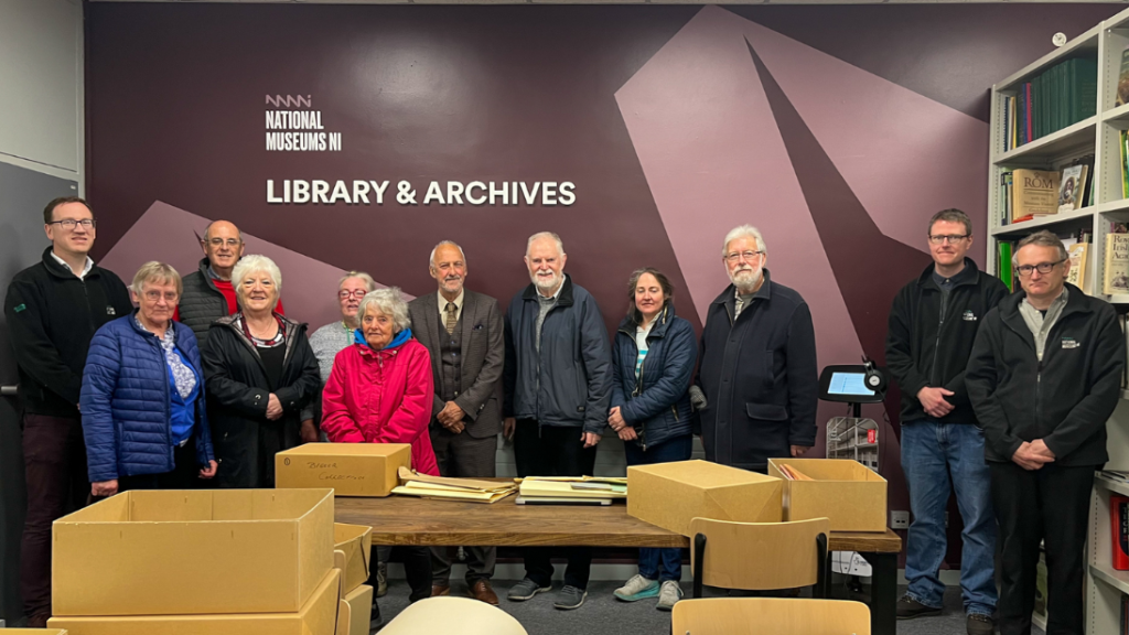A group of people stand in a library. The background wall is purple with big white letters reading ' National Museums NI Library & Archives'.