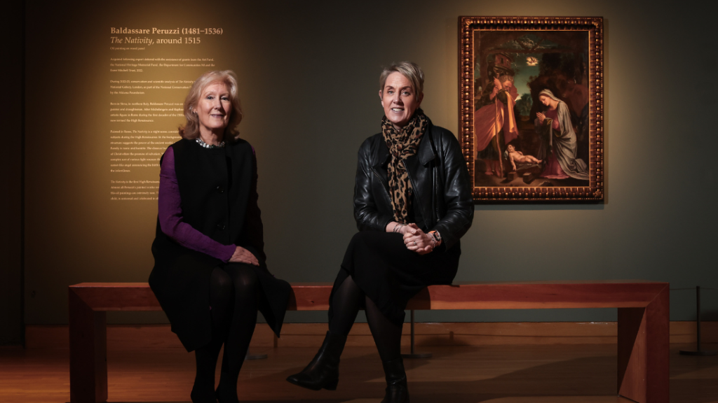 Two women sitting on a bench inside an art gallery at the Ulster Museum with a Peruzzi painting in the background and white writing on the wall.