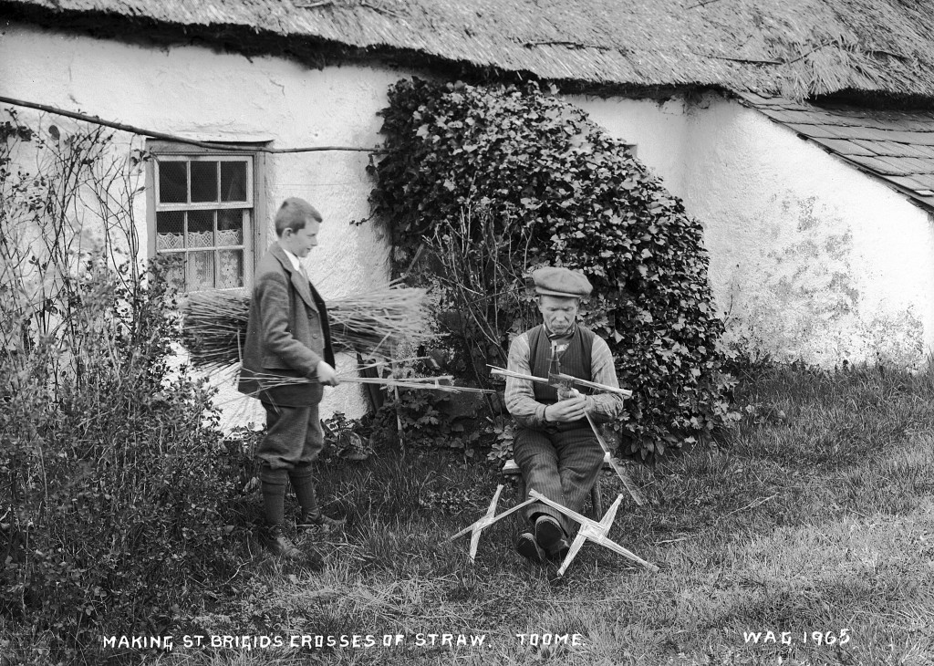 A black and white image of two men making St Brigid's crosses outside of a cottage.