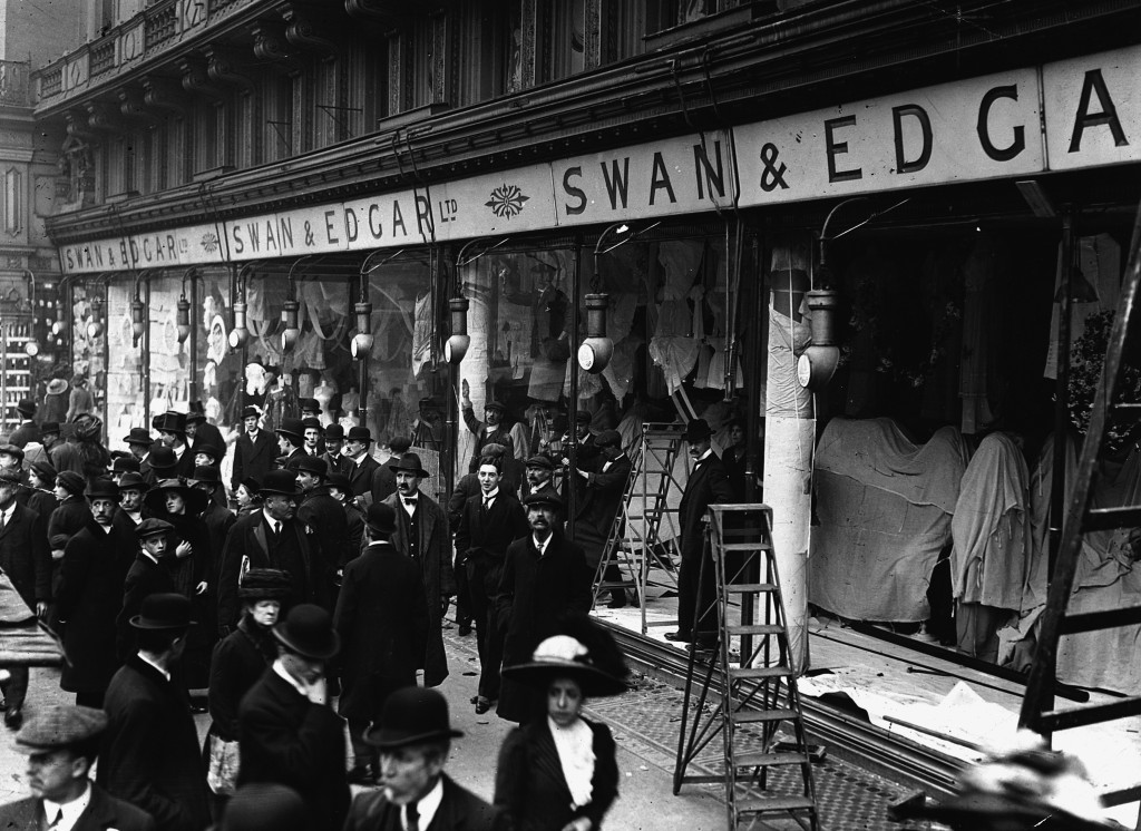 A group of people stand outside of a shop in a black and white photgraph. Some of the windows, which are floor to ceiling, appear to be missing.