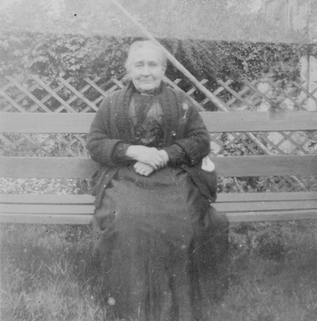A smiling older woman looks at the camera in a black and white photograph. She is sitting outdoors on a bench wearing a long dark dress,