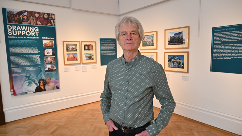 A man with grey hair, dressed in a shirt, hands in pockets standing inside the Belfast Room of Ulster Museum, at the reveal of a new murals photography exhibition. Framed photographs are on white walls behind him as well as a turquoise wall graphic that says 'Drawing Support'.