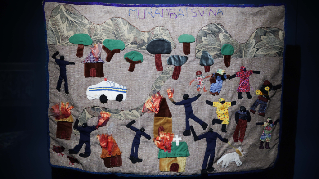 A colourful arpillera, on a grey linen background, made by a group of girls in Killarney, Zimbabwe in 2012. It shows the destruction of their informal settlement in May 2005 during the government led ’Operation Murambatsvina’ (get rid of the filth) in which an estimated 500,000 people were evicted and displaced in the space of a month.