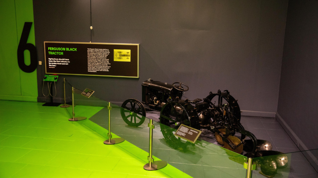 A black tractor is displayed behind glass railings. The wall and part of the floor is painted a dark navy which joins another section of floor painted bright green. There is a large number 6 on the wall beside a backlit sign with text on it about the tractor on display.