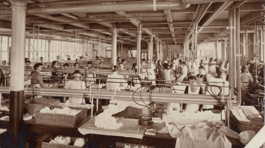 A black and white image of a workroom with many women and girls working at sewing.