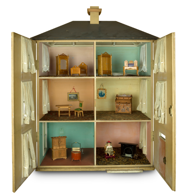 A tall yellow dolls' house with its front opened so we can see each room. There are six rooms, some with a dresser and bed, one with a writing desk, and a living room with a doll sitting in a chair.