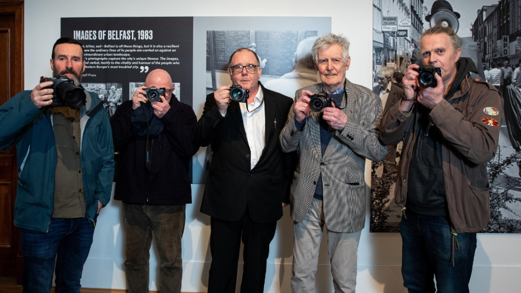 Five photographers lined up in a row holding their cameras, posing as if they're taking photos